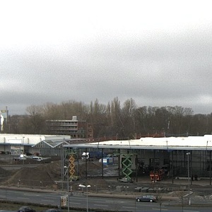 Blick auf eine Baustelle mit mehreren Baugeräten und Maschinen. Im Hintergrund sind Bäume und ein mehrstöckiges Gebäude zu sehen. Der Himmel ist bewölkt und die Umgebung wirkt städtisch. Einige Autos fahren auf der Straße entlang.