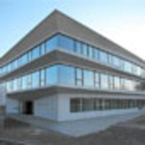 Eine moderne, dreigeschossige здание mit großen Glasfenstern und einem minimalistischen Design. Das Gebäude hat eine helle Fassade und befindet sich in einem klaren Himmelshintergrund. Der Eingang ist auf der linken Seite sichtbar.