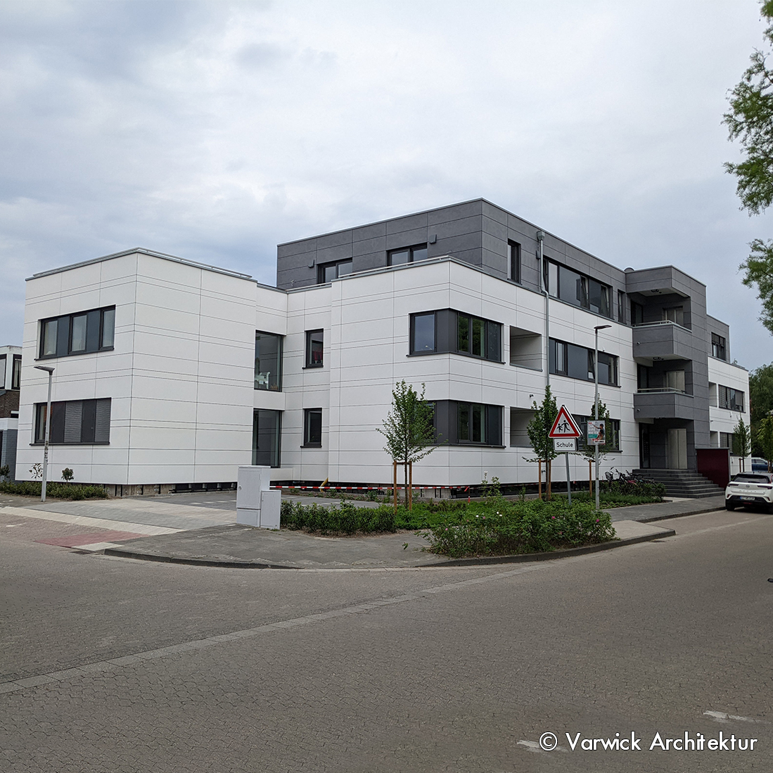 Moderner Wohngebäudekomplex in hellen Farben, mit mehreren Etagen und großen Fenstern. Vor dem Gebäude sind kleine Bäume und ein Weg platziert. Der Himmel ist bewölkt und es sind einige Fahrzeuge in der Nähe geparkt.