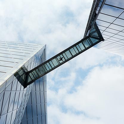 Die Bildbeschreibung zeigt eine moderne Architektur mit zwei hohen Gebäuden, die durch einen gläsernen Verbindungsgang miteinander verbunden sind. Der Himmel ist teilweise bewölkt und die Oberflächen der Gebäude reflektieren das Licht.