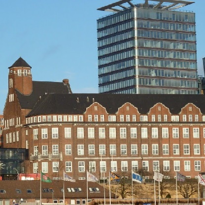 Das Bild zeigt zwei Gebäude: ein historisches, kubisches Backsteingebäude mit vielen Fenstern und ein modernes, gläsernes Hochhaus. Vor den Gebäuden sind mehrere Fahnen zu sehen, die im Wind wehen. Der Himmel ist klar und blau.