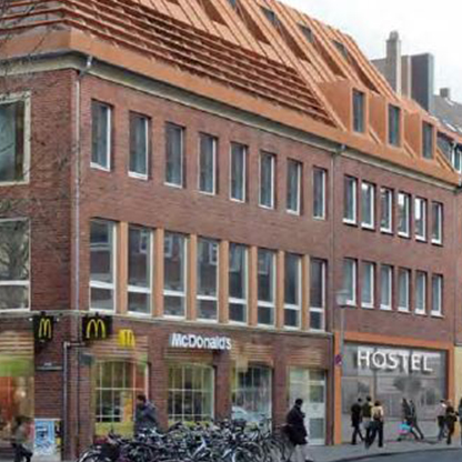 Eine moderne Backsteinfassade mit einem Hostel und einem McDonald's im Erdgeschoss. Das Gebäude hat große Fenster und ein auffälliges, rot eingedecktes Dach. Vor dem Haus sind viele Fahrräder abgestellt, und Menschen gehen auf dem Bürgersteig.