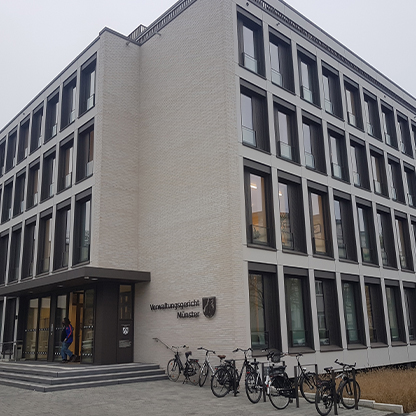 Ein modernes, mehrstöckiges Gebäude mit großen Fenstern und einem klaren, funktionalen Design. Vor dem Eingangsbereich stehen mehrere Fahrräder. An der Fassade ist das Schild "Verwaltungsgericht Münster" angebracht. Der Himmel ist bewölkt.