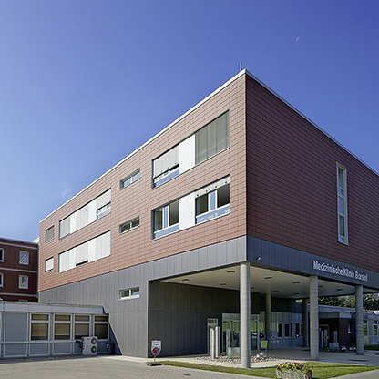 Moderne medizinische Einrichtung mit einem dreigeschossigen Gebäude. Die Fassade ist in Braun- und Beigetönen gehalten und hat große Fenster. Im Vordergrund ist der Eingang mit einem überdachten Bereich zu sehen. Blauer Himmel und einige Pflanzen im Vordergrund.
