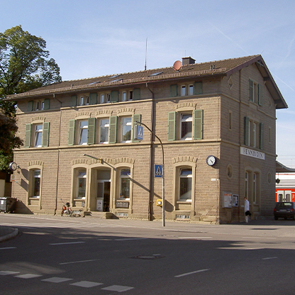 Historisches Gebäude an einer Straßenecke mit grünem Fensterladen und Uhr an der Wand. Es handelt sich offenbar um einen Bahnhof oder ein ähnliches öffentliches Gebäude. Bäume sind im Hintergrund sichtbar, und einige Personen sind in der Nähe unterwegs.