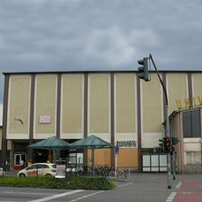 Ein Gebäude mit einer großen, hellen Fassade und mehreren Fenstern. Vor dem Gebäude befinden sich zwei transparente Überdachungen und ein Verkehrssignal. Der Himmel ist bewölkt, und es gibt einige Fahrräder in der Nähe.