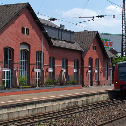 Ein rotes Ziegelgebäude, vermutlich ein Bahnhofsgebäude, mit großen Fenstern und Sonnenschirmen im Außenbereich. Auf dem Gleis steht ein moderner Zug. Im Hintergrund ist ein Werbeschild für ein Busunternehmen sichtbar, bei klarem Himmel.