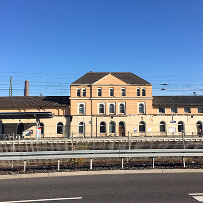 Ein Bahnhofgebäude mit einer hellbeigen Fassade und mehreren Fenstern ist im Vordergrund zu sehen. Der Himmel ist klar und blau. Im Hintergrund sind Oberleitungen und Züge zu erkennen, die für den Schienenverkehr typisch sind.