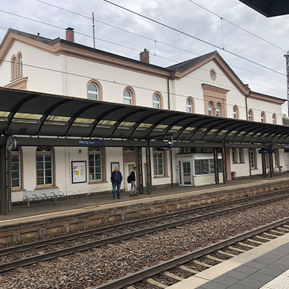 Bahnhof mit einem weißen Gebäude, das ein längliches Vordach hat. Zwei Personen stehen auf dem Bahnsteig. Gleise verlaufen vor dem Gebäude. Über den Gleisen sind Informationsschilder und eine Haltestelle sichtbar. Der Himmel ist bewölkt.