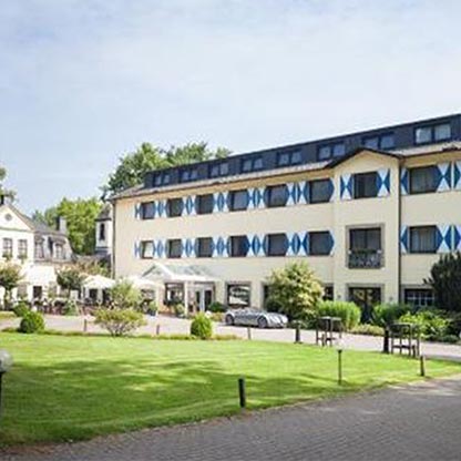 Eine Ansicht eines Hotels mit drei Etagen, das eine moderne Architektur aufweist. Die Fassade ist in hellem Gelb mit blauen Fensterläden gehalten. Im Vordergrund erstreckt sich ein gepflegter grüner Rasen mit Sträuchern und einem Parkplatz.