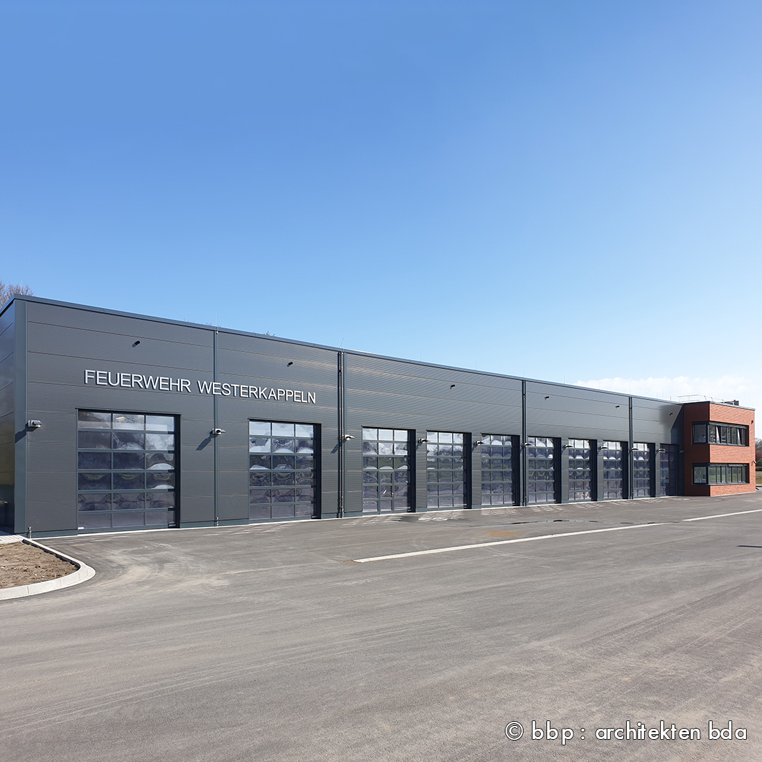 Moderne Feuerwache in Westerkappeln mit einer großen, verglasten Front. Die Fassade ist grau und schließt mehrere Rolltore für Feuerwehrfahrzeuge ein. Im Hintergrund ist ein blauer Himmel sichtbar.