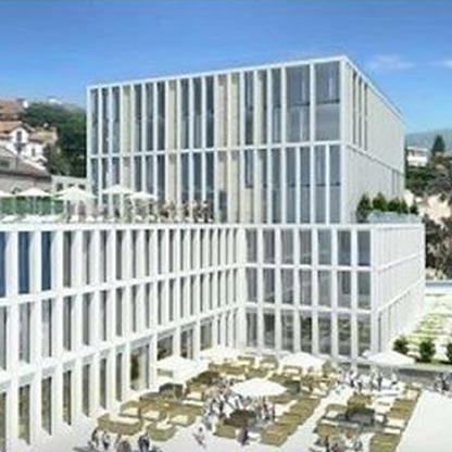 Moderne Architektur eines Gebäudes mit mehreren Stockwerken. Die Fassade ist mit großen Glasflächen und weißen Wänden gestaltet. Im Vordergrund befinden sich Terrassen mit Sitzgelegenheiten und Sonnenschirmen, umgeben von Menschen. Der Himmel ist klar und blau.