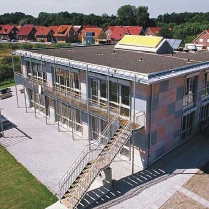 Modernes zweigeschossiges Gebäude mit großflächigen Fensterfronten, einer Treppe an der Seite und farbigen Fassaden. Im Hintergrund sind Reihenhäuser mit roten Dächern sichtbar. Der Außenbereich ist gepflastert und von Rasen umgeben.