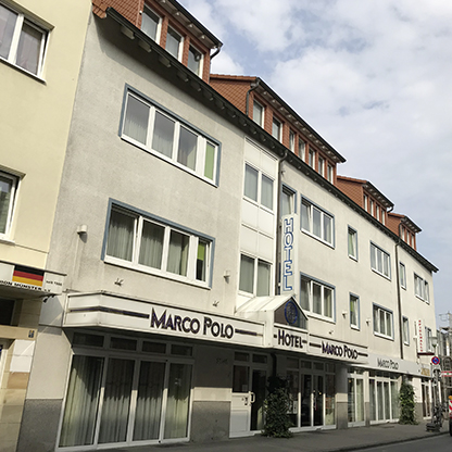 Ein mehrstöckiges Gebäude mit der Beschriftung "Hotel Marco Polo" an der Fassade. Die Fenster sind überwiegend hell und der Eingang ist mit einer Veranda ausgestattet. Die Architektur ist modern und schlicht, mit einem grauen und weißen Farbschema.