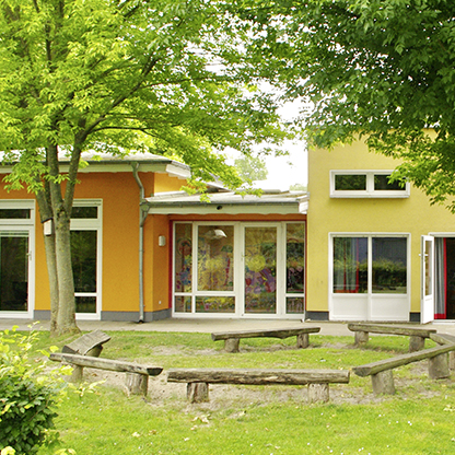 Ein modernes, einseitig gefärbtes Gebäude mit großen Fenstern, umgeben von Bäumen und Grünflächen. Vor dem Gebäude bildet ein kreisförmiger Holzstamm-Sitzplatz eine einladende Versammlungszone im Freien. Der Himmel ist klar und sonnig.