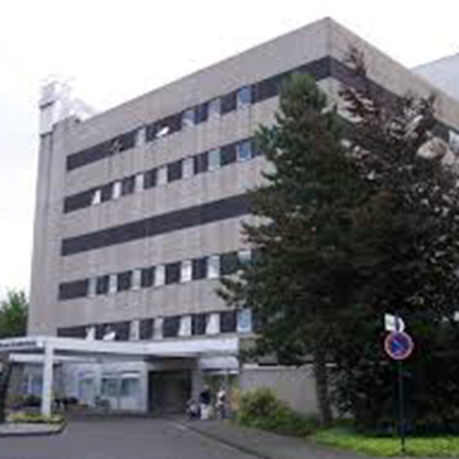Modernes Gebäude mit sechs Stockwerken, grauer Fassade und mehreren Fenstern. Im Vordergrund befindet sich der Eingang mit einem überdachten Bereich. Umgeben von Bäumen und Grünflächen.