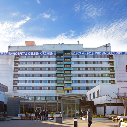 Das Bild zeigt die Fassade des Marienhospitals in Gelsenkirchen. An der Vorderseite sind die Jahreszahlen 1869 und 2019 sowie der Slogan "WIR SIND GESUNDHEIT" sichtbar. Im Vordergrund sind einige Menschen zu sehen, die sich auf dem Gelände bewegen.