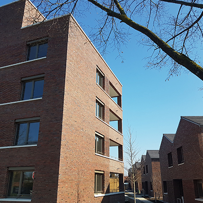 Moderne Ziegelgebäude in einer städtischen Umgebung, unter einem klaren blauen Himmel. Links steht ein höheres Gebäude mit mehreren Fenstern, während rechts ein niedrigeres, angrenzendes Gebäude zu sehen ist. Äste eines Baumes rahmen die Szene ein.
