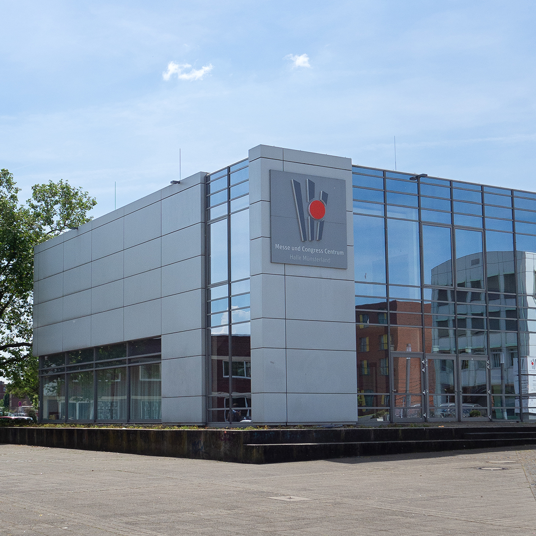Das Bild zeigt die moderne Fassade eines Gebäudes, das als "Messe- und Kongresszentrum" bezeichnet ist. Es hat große Glasfenster und eine auffällige Gestaltung, umgeben von einem gepflasterten Platz und einigen Bäumen im Hintergrund unter einem klaren Himmel.