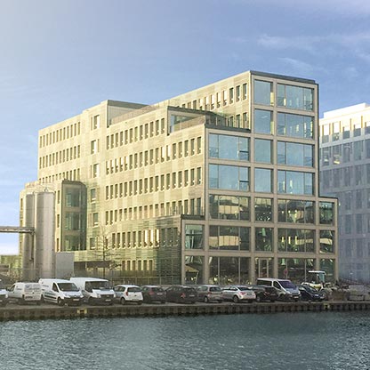 Moderner Bürogebäude am Wasser mit großen Fensterfronten und mehreren Etagen. Umgeben von Autos auf einem Parkplatz und einem weiteren Gebäude im Hintergrund. Der Himmel ist klar und sonnig.