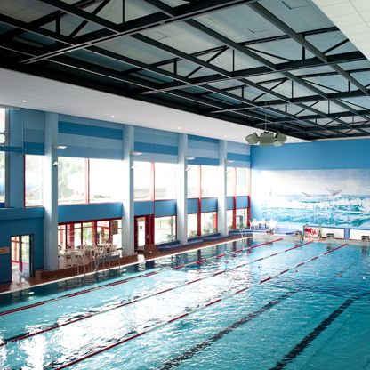 Ein Innenbereich eines Schwimmbades mit einem Becken, das von großen Fenstern umgeben ist. An einer Wand ist ein Wandbild mit einer Meeresszene zu sehen. Die Beleuchtung ist modern und der Raum wirkt hell und einladend.