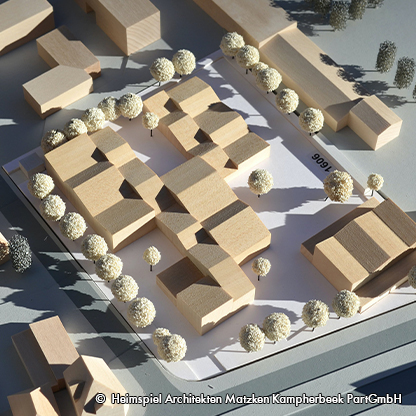 Modell einer Architekturplanung mit mehreren holzfarbenen Gebäuden, umgeben von kleinen, runden Bäumen. Die Struktur ist auf einem weißen Untergrund platziert. Die Anordnung der Gebäude ist klar und geometrisch.