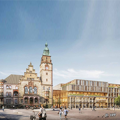 Eine Ansicht eines Stadtplatzes mit einem historischen Gebäude und einem modernen Bau. Im Vordergrund gehen Personen, und im Hintergrund ist ein Turm mit einer Uhr zu sehen. Der Himmel ist klar und blau.