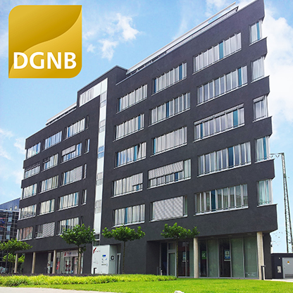 Ein modernes, mehrstöckiges Bürogebäude mit dunkler Fassade und großen Fenstern. Im Vordergrund sind einige kleine Bäume und ein Rasen zu sehen. Oben links befindet sich ein Logo mit den Buchstaben "DGNB". Der Himmel ist blau mit einigen Wolken.