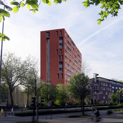 Ein modernes, mehrstöckiges Gebäude mit rot-orangefarbener Fassade steht im Vordergrund, umgeben von Bäumen und Grünanlagen. Im Hintergrund sind weitere niedrigere Gebäude sichtbar. Der Himmel ist klar mit einigen Wolken.
