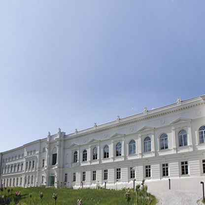 Die Abbildung zeigt eine historische, weiße Gebäudeansicht mit mehreren Fenstern und architektonischen Details unter einem klaren blauen Himmel. Eine grüne Wiese mit einigen Blumen befindet sich im Vordergrund.