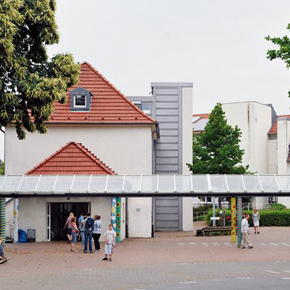 Ein Gebäude mit rotem Dach und moderner Fassade, umgeben von Bäumen. Vor dem Eingang stehen mehrere Personen. Der Bereich ist gepflastert, und es gibt eine Überdachung mit bunten Säulen. Die Atmosphäre wirkt einladend und freundlich.