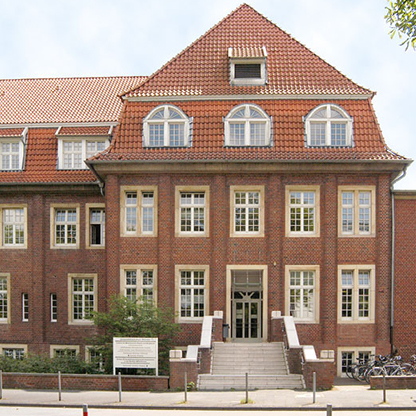 Ein dreistöckiges Backsteingebäude mit rotem Satteldach. Die Fassade hat große Fenster und eine breite Treppe, die zum Eingang führt. Vor dem Gebäude sind einige Fahrräder abgestellt, und es sind Bäume sowie eine Informationsplatte sichtbar.