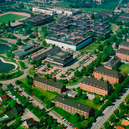 Luftaufnahme eines weitläufigen Betriebsgeländes mit mehreren großen Gebäuden, Parkplätzen und umliegenden Grünflächen. Ein Teich und Straßen sind ebenfalls zu sehen, umgeben von Wohngebäuden.