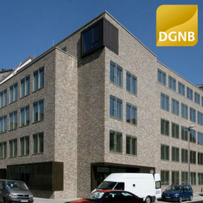 Eine moderne, mehrstöckige Gebäudefassade aus hellen Klinkern mit großen Fenstern. Im Vordergrund sind einige Autos geparkt. Oben rechts ist ein Logo der DGNB (Deutsche Gesellschaft für Nachhaltiges Bauen) zu sehen.