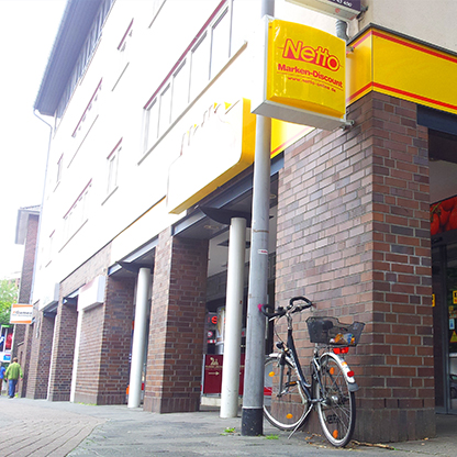 Ein Fahrrad steht an einem Laternenpfahl vor einem Geschäft mit dem Schild "Netto Marken-Discount". Das Gebäude ist aus roten Ziegeln und hat große Fenster. Menschen sind im Hintergrund zu sehen.