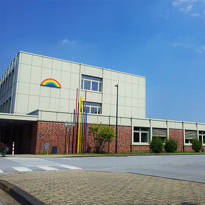 Ein modernes Gebäude mit einer grauen Fassade und einem Ziegelsockel. An der Wand ist ein farbenfrohes Regenbogen-Symbol angebracht. Vor dem Gebäude stehen mehrere farbige Stäbe. Der Himmel ist klar und blau, und es sind einige Pflanzen und Bäume sichtbar.
