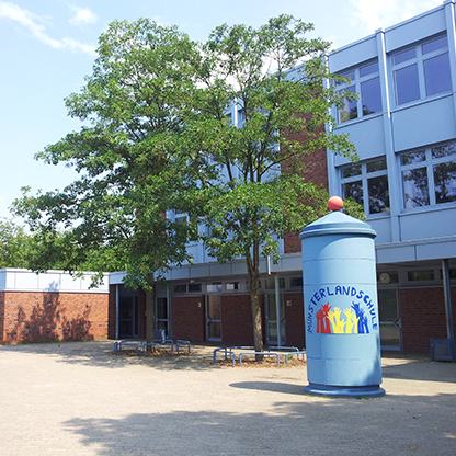 Das Bild zeigt den Außenbereich einer Schule mit einem großen blauen Zylinder, der das Logo der "Münsterland-Schule" trägt. Im Hintergrund stehen Fenster und ein Baum ist sichtbar. Der Boden ist gepflastert und einige Bänke sind aufgestellt.
