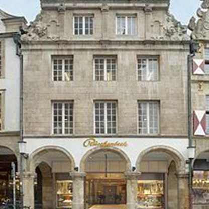 Die Fassade eines historischen Gebäudes mit mehreren Fenstern und einem gewölbten Eingangsbereich. Der Name des Geschäfts "Rüschenbeck" ist sichtbar. Über dem Eingangsbereich befinden sich architektonische Verzierungen.