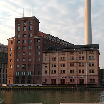 Ein historischer Backsteinbau mit mehreren Etagen an einem Gewässer. Das Gebäude trägt die Aufschrift "WOLFGANG BORCHERT THEATER" und hat große Fenster. Im Hintergrund ist ein hoher Schornstein und eine bewölkte Himmelsszenerie sichtbar.