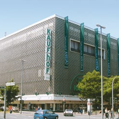 Eine moderne Kaufhausfassade mit dem Schriftzug "KAUFHOF" in großen Buchstaben. Das Gebäude hat mehrere Fenster und ist von Bäumen und einer Straße umgeben. Es gibt Verkehrsschilder und Autos im Vordergrund.