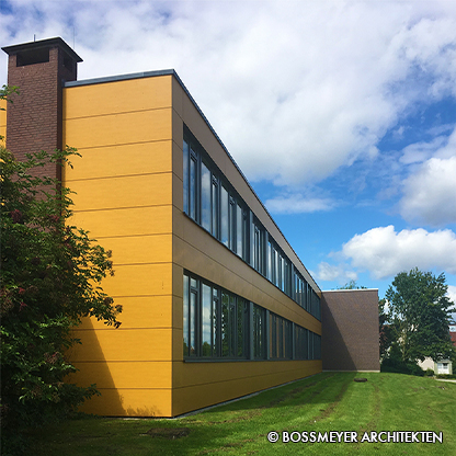 Das Bild zeigt ein modernes Gebäude mit einer gelben Fassadenverkleidung und großen Fenstern. Auf der linken Seite befindet sich ein Kamin, und im Hintergrund sind Bäume und Gras zu sehen. Der Himmel ist teilweise bewölkt.