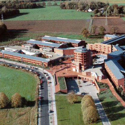 Luftaufnahme eines modernen Gebäudekomplexes, umgeben von Grünflächen und Feldern. Die Architektur zeichnet sich durch rechteckige Formen und eine zentrale Turmstruktur aus. Eine Straße führt entlang des Gebäudes, und mehrere Fahrzeuge sind sichtbar.