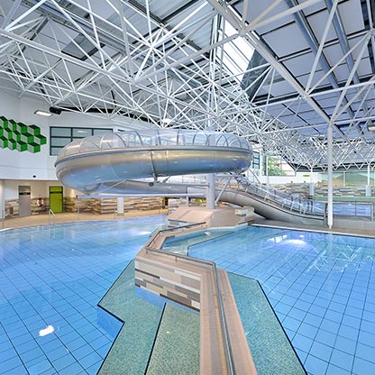Ein modernes Schwimmbad mit einer blauen Wasseroberfläche und einer großen Rutsche. Das Gebäude hat hohe, lichtdurchflutete Decken mit einem Metallgerüst und grün gestalteten Wandpartien. Die Rutsche führt in das Becken, das flach und einladend wirkt.