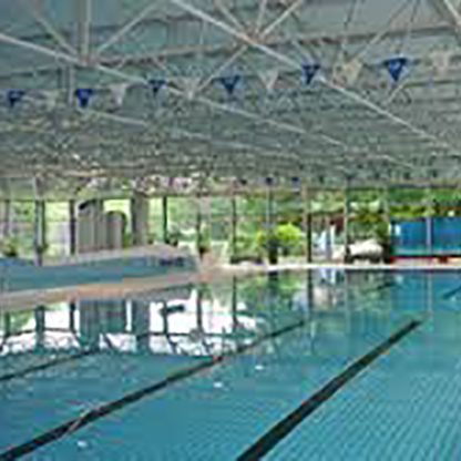 Ein Innenpool mit klarem Wasser und mehreren Schwimmbahnen. Das Dach ist aus Metall mit sichtbaren Balken, und es gibt blaue Wimpel, die zu den Seiten hängen. Durch große Fenster ist viel Tageslicht zu sehen, und grüne Bäume sind im Hintergrund erkennbar.