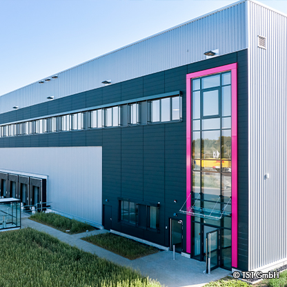 Moderne Lagerhalle mit einer markanten Eingangstür in pink und großen Fenstern. Die Fassade ist schmal und hat eine Kombination aus dunklen und hellen Paneelen. Vor dem Gebäude sind Rasenflächen, die zu einem gepflasterten Weg führen.