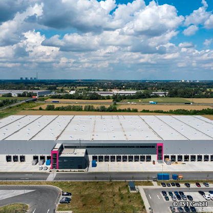 Luftaufnahme eines großen Lagerhauses mit flacher Dachkonstruktion. Das Gebäude hat eine graue Fassade, mehrere Andockstationen und eine moderne Eingangszone in Pink. Um das Lager sind Parkplätze und eine grüne Landschaft zu sehen, unter einem bewölkten Himmel.