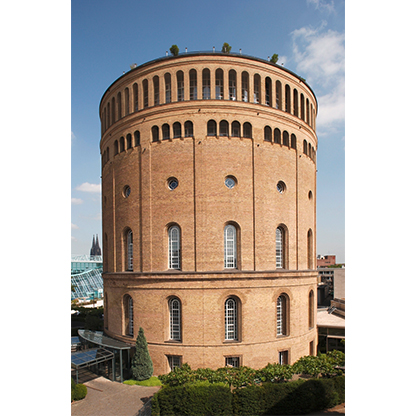 Runder, backsteingelber Wasserturm mit großen Fenstern und einem leicht gewölbten Dach. Umgeben von grünen Pflanzen und Bäumen, unter blauen Himmel mit einigen Wolken. Im Hintergrund sind moderne Gebäude und Türme sichtbar.