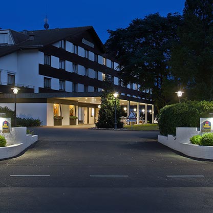 Eingang zu einem Hotelgebäude bei Nacht, beleuchtet mit Straßenlaternen. Der Eingang ist von grünen Pflanzen umgeben, und im Hintergrund sind Fenster mit warmem Licht zu sehen. Das Gesamtbild vermittelt eine einladende Atmosphäre.