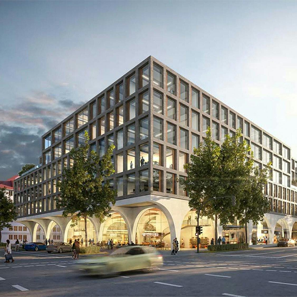 Moderne Architektur eines mehrstöckigen Gebäudes mit klaren Linien und großen Fensterflächen. Das Gebäude steht an einer Straßenecke, umgeben von Bäumen. Im Vordergrund sind Autos und Fußgänger zu sehen, während der Himmel in den Abendstunden beleuchtet ist.