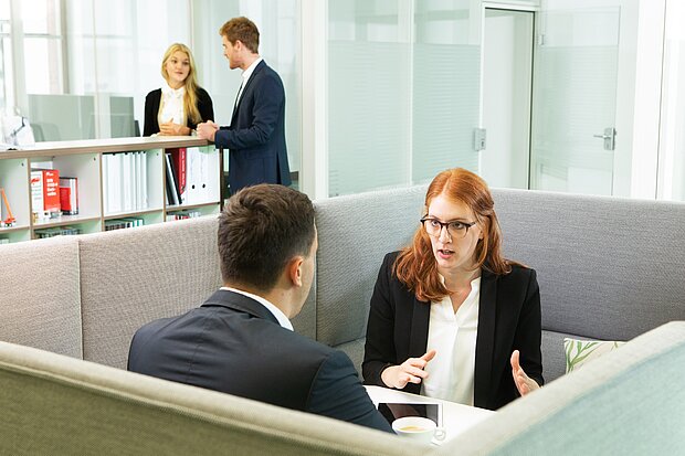 Zwei Personen in Anzügen führen ein Gespräch in einem modernen Büro-Setup, während eine dritte Person im Hintergrund an einem Tisch steht und mit einer weiteren Person spricht. Die Gesprächspartner scheinen engagiert und diskutieren lebhaft.